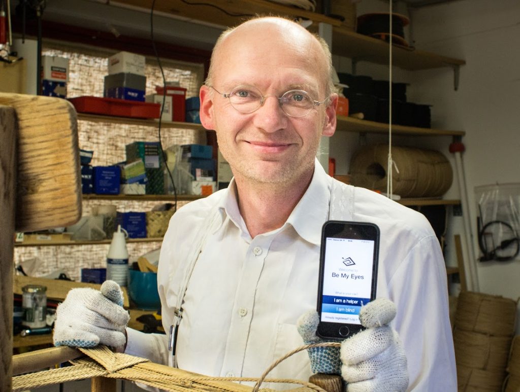 foto do criador do aplicativo Be my Eyes em uma oficina de artesanato. Ele é um homem calvo, com (poucos) cabe,os grisalhos e barba por fazer. Usa óculos e tem olhos claros. Está sorrindo para a foto e segura um celular com a página inicial do app. 