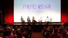 Imagem do evento realizado anteriormente, mostrando palestrantes em um palco, do ponto de vista da platéia.