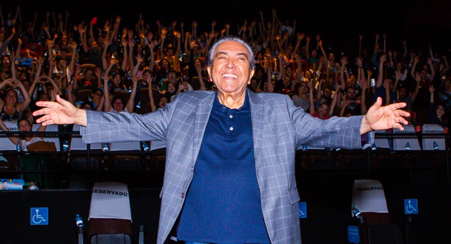 Maurício de Souza, criador da turma da Mônica, posa sorrindo de braços abertos, à frente da platéia do cinema, na pré-estreia de Turma da Mônica Jovem