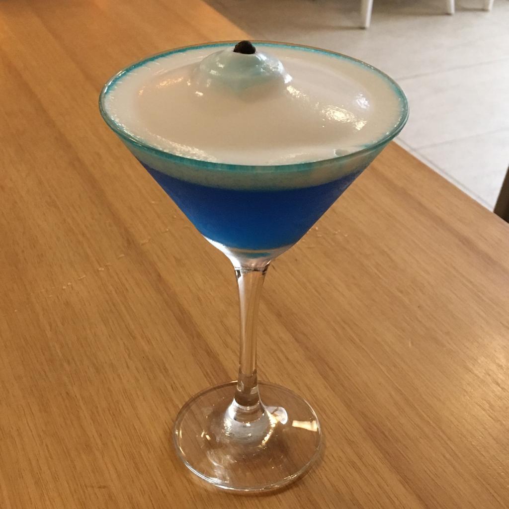 Imagem de taça com o drink "olho grego": uma bebida azul, com espuma branca em cima e um pontinho preto de uma semente no centro.