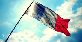 Imagem da bandeira da França tremulando em um mastro, com o céu azul com algumas nuvens brancas.