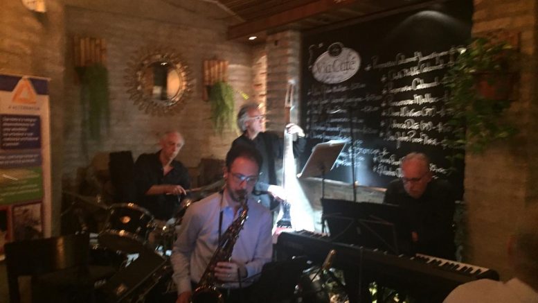 Quarteto musical tocando em um local com pouca luz, em primeiro plano o saxofonista tocanndo sentado.