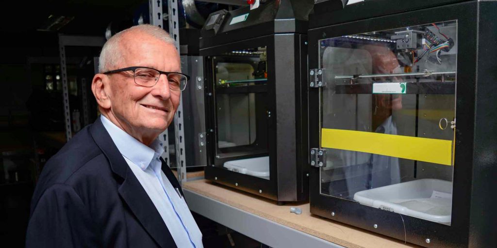 Imagem do fundador da empresa, um senhor de cabelos brancos, sorrindo para a câmera à frente de uma impressora 3D.