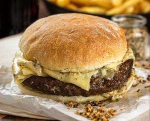 Imagem de um hamburguer em primeiro plano, com pão e queijo. Atrás dele se percebe uma porção de batatas fritas. 