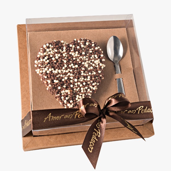 Coração de chocolate e colher de alumínio, embalados em uma caixa com tampo transparente  fechada com laço de fita. 