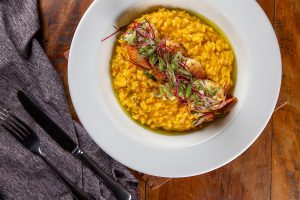 Imagem mostra um prato branco com risoto de abóbora e camarões dispostos sobre o risoto como uma faixa. Sobre eles algumas verduras.