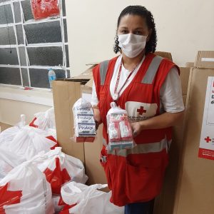 mulher com colete vermelho, vestindo máscara facial, posa ao lado de sacos e caixas com kits de higiene. Ela tem um kit em cada mão.