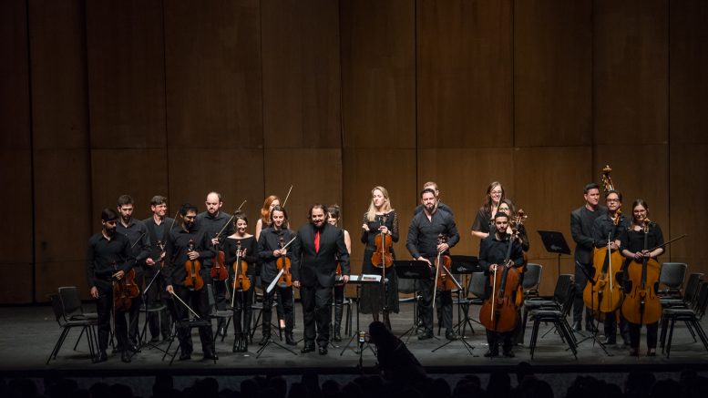 imagem de músicos de uma orquestra no palco, todos em pé, vistos de longe, no momento de agrdecimento.