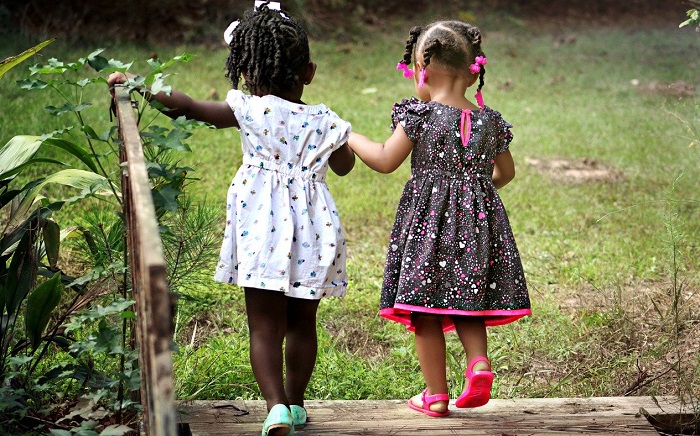 imagem mostra duas crianças pequenas, de costas caminhando de mãos dadas, elas estao com vestidos floridos