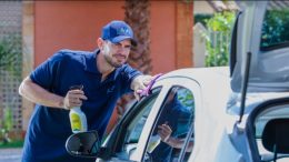 imagem mostra homem passando produto de limpeza em um carro. Ele veste uniforme da lavô, com camiseta e boné azuis e está sorrindo..