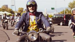uma rua com diversos motociclistas, em primeiro plano, um homem de paletó azul, colete amarelo estampado e gravata borboleta vermelha pilota uma moto sorrindo, ele usa luvas de couro, um capacete antigo e óculos escuros.