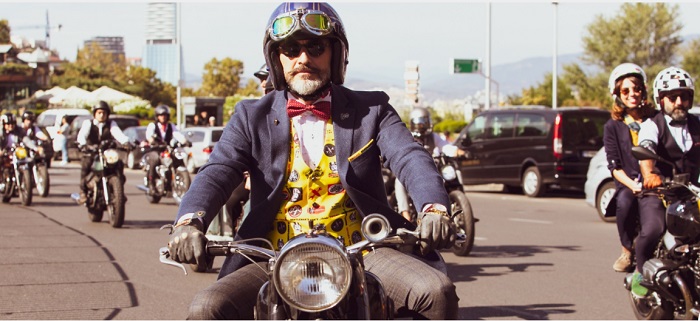 uma rua com diversos motociclistas, em primeiro plano, um homem de paletó azul, colete amarelo estampado e gravata borboleta vermelha pilota uma moto sorrindo, ele usa luvas de couro, um capacete antigo e óculos escuros.