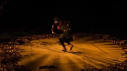imagem mostra cena de teatro, em que em um local escuro, tendendo ao ocre, um homem parece realizar uma dança dentro de um circulo no chão delimitado por folhas secas
