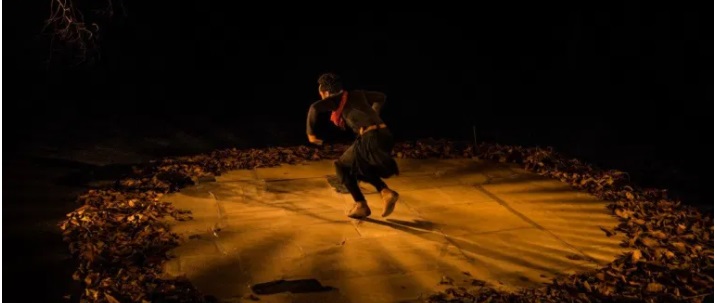 imagem mostra cena de teatro, em que em um local escuro, tendendo ao ocre, um homem parece realizar uma dança dentro de um circulo no chão delimitado por folhas secas
