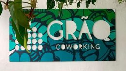 imagem da placa do Grão coworking, estampada em tons de azul e verde. ela é mostrada em uma parede branca, com algumas folhagens sobre ela.