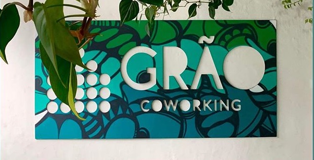 imagem da placa do Grão coworking, estampada em tons de azul e verde. ela é mostrada em uma parede branca, com algumas folhagens sobre ela.
