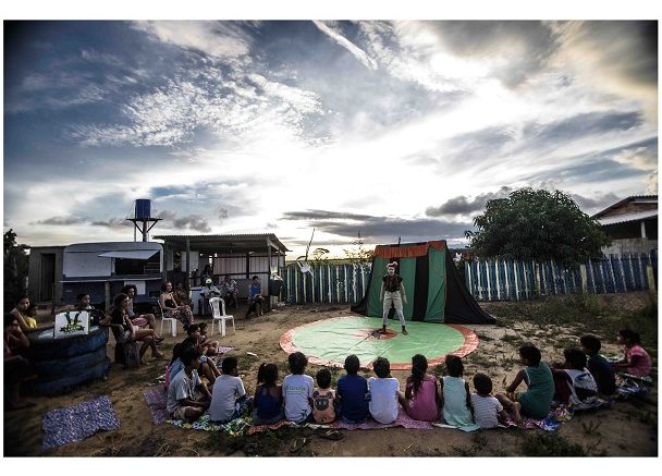 imagem de um grupo sentdo em roda, de costas para a foto, assistindo um espetáculo no chão, ao ar livre.