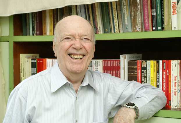 autor sorri, com um dos braços apoiados em uma estante cheia de livros