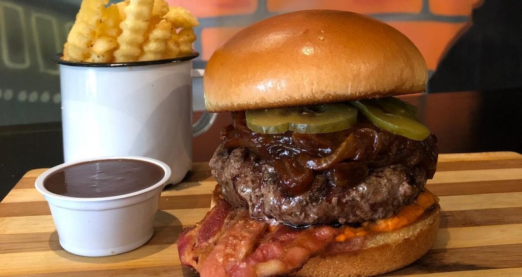 Imagem de um hamburguer alto, com um potinho de molho e uma caneca com fritas. O Feline, participante do Burguer Fest