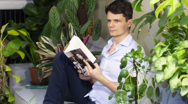 ator sentado no meio de várias plantas, com um livro de Clarice na mao.