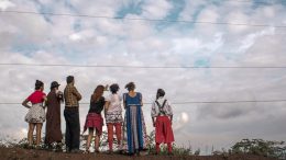 Se vê ao longe um grupo de palhaços de costas, sobre um chão de terra e sob um céu azul com algumas núvens