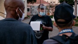dois moradores de rua, de costas, usando máscaras, conversam com um monitor do projeto, que usa máscara e face shield e segura uma prancheta.