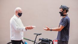 O pai Antonio vestido uma doma branca, entrega um Torta do Pai para o filho Diego, que está com capacete de bike. ambos vestem máscara.