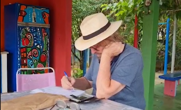 imagem de Carlos Bracher sentado á mesa, pintando. ele usa chapéu panama e está com o rosto apoiado em uma mão, atrás dele é possível ver algumas plantas, sobre a mesa muitos papeis e seu celular.
