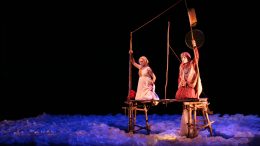 cena da peça Saudade em Terras dagua, dois atores em pé em um barco estilizado, o palco é escuro e há uma luz aul no chão