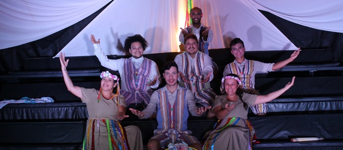 artistas do 'ensaio a paixão de cristo em cordel" sentados em uma arquibancada, erguendo os braços como um cumprimento para o público.