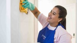 imagem de uma mulher sorrindo ao passar um pano amarelo, suportamente tirando restos de cola da parede. ela usa uma luva plastica azul clara e um avental azul royal.