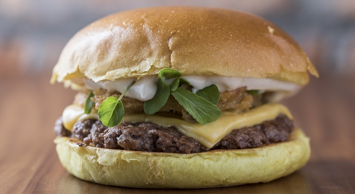 imagem de um sanduiche laçado no dia do hamburguer, ele é visto de lado, com pão de hambuerguer, queijo rucula e um molho branco.