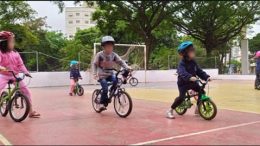 imagem de três crianças em pequenas bikes em primeiro plano e outras atrás, todas em uma quadra durante atvidade do Aromeiazero