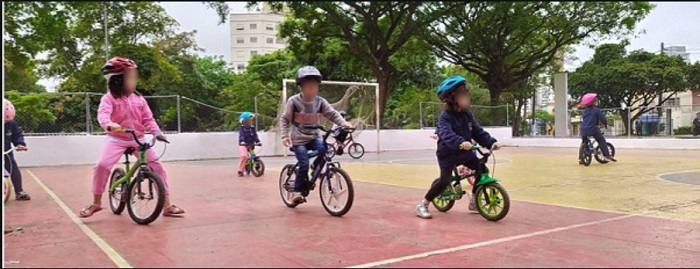 imagem de três crianças em pequenas bikes em primeiro plano e outras atrás, todas em uma quadra durante atvidade do Aromeiazero
