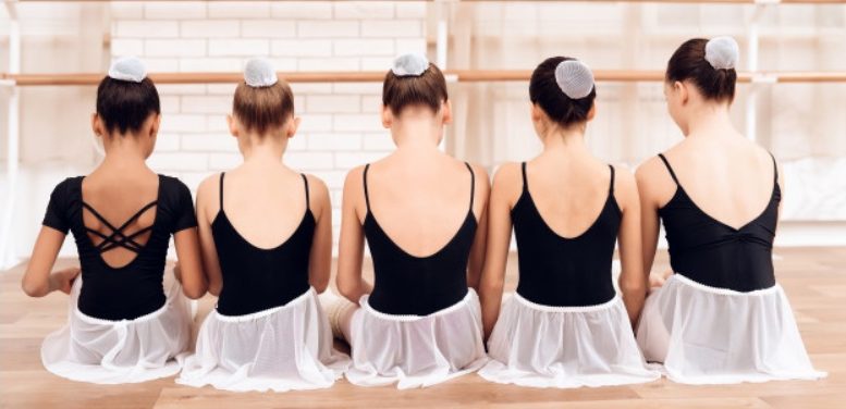 cinco meninas bilarinas sentadas de costas. todas vestem collant preto de dança e saia de bailarina rosa., alem de ter cabeo preso em coque com redinha.