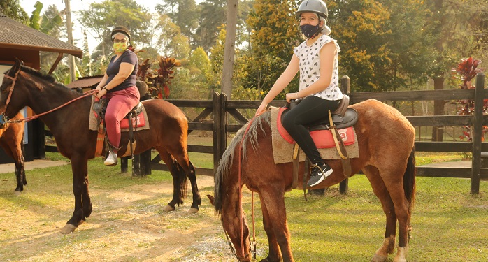 imagem de duas jovens com máscara, montadas em cavalos em uma área verde,