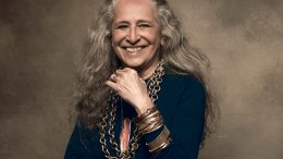 Bethânia posa, em plano americano, sorrindo, com blusa azul, colares e pulseiras de metal, ela tem uma das mãos junto ao queixo.