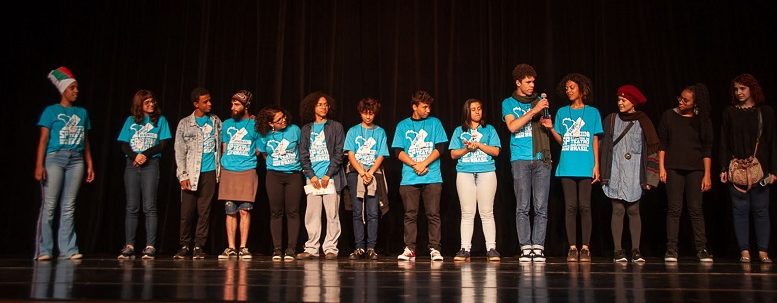 imagem de um palco com diversos adolescentes lado a lado sobre ele. Todos vestem uma camiseta azul e calças em estilos variados.