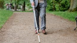 imagem de uma pessoa cega andando em uma alameda de um parque com auxílio de uma bengala, no caso branca com vermelho.
