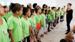 imagem de crinças do projeto escola de trovadores perfiladas, todas com camisetas veerde claro há uma professora em frente, estindo preto, ela parece cantar