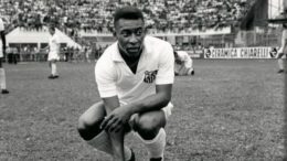 imagem em preto e branco do jogador Pelé, ainda jovem, com uniforme do Santos, agaichado no gramado.