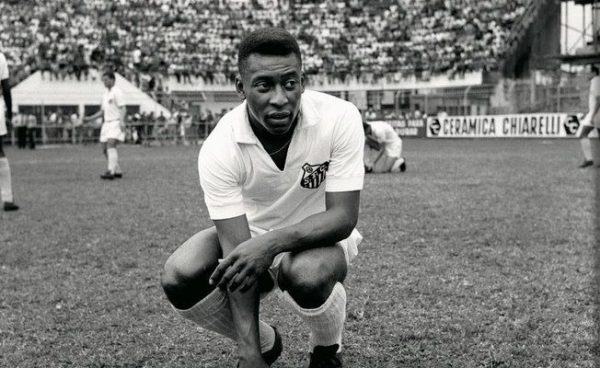 imagem em preto e branco do jogador Pelé, ainda jovem, com uniforme do Santos, agaichado no gramado.