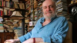 imagem do compositor Aldir Blanc sentado em uma poltrona à frente de estantes com diversos livros. Ele está com camisa azul de manga longa e sorri para a foto.