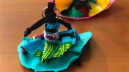 imagm de uma boneca abayomi, a pequena boneca de tecido é preta, tem vestido azul com detalhes em estamparia etnica africana e colocada sentada sobre uma mesa de madeira.