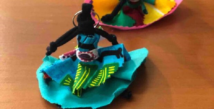 imagm de uma boneca abayomi, a pequena boneca de tecido é preta, tem vestido azul com detalhes em estamparia etnica africana e colocada sentada sobre uma mesa de madeira.