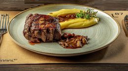 jogo americano com um prato de carne e purê, e talheres ao lado.