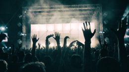 imagem de um show de rock, se vê pela perspctiva do publico que está no fundo da plateia, um palco iluminado e vários braços erguidos.