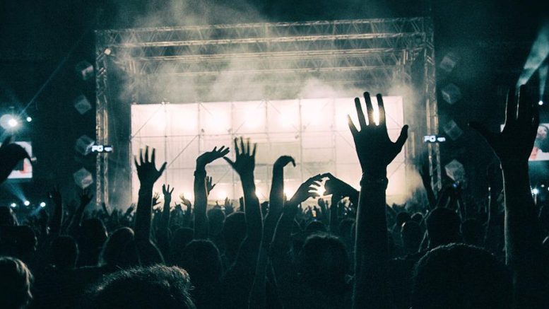 imagem de um show de rock, se vê pela perspctiva do publico que está no fundo da plateia, um palco iluminado e vários braços erguidos.