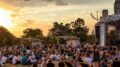 imagemde pessoas sentads no chão de um parte, um palco coberto e o por do sol