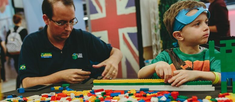 garoto fantasiado de super heroi, com a máscara levantada, brinca em uma mesa cheia de peças de lego, ao lado um adulto também monta pecinhas